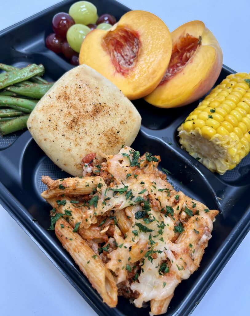 School lunch tray with peaches, corn on the cobb, grapes, green beans, Beef Ragout with Penne pasta and a roll.