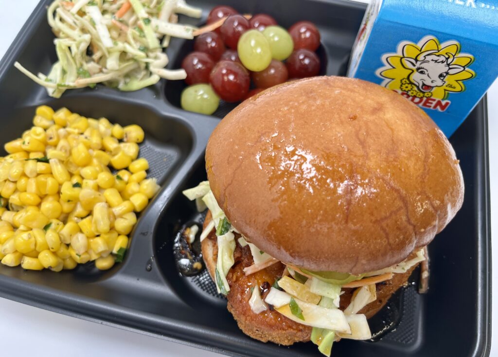 School lunch tray with chicken sandwich, slaw, grapes, steamed corn, and milk.