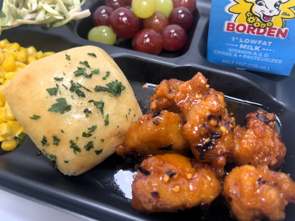 School lunch tray with Hot Honey Boneless Wings, roll, corn, slaw, grapes and milk.