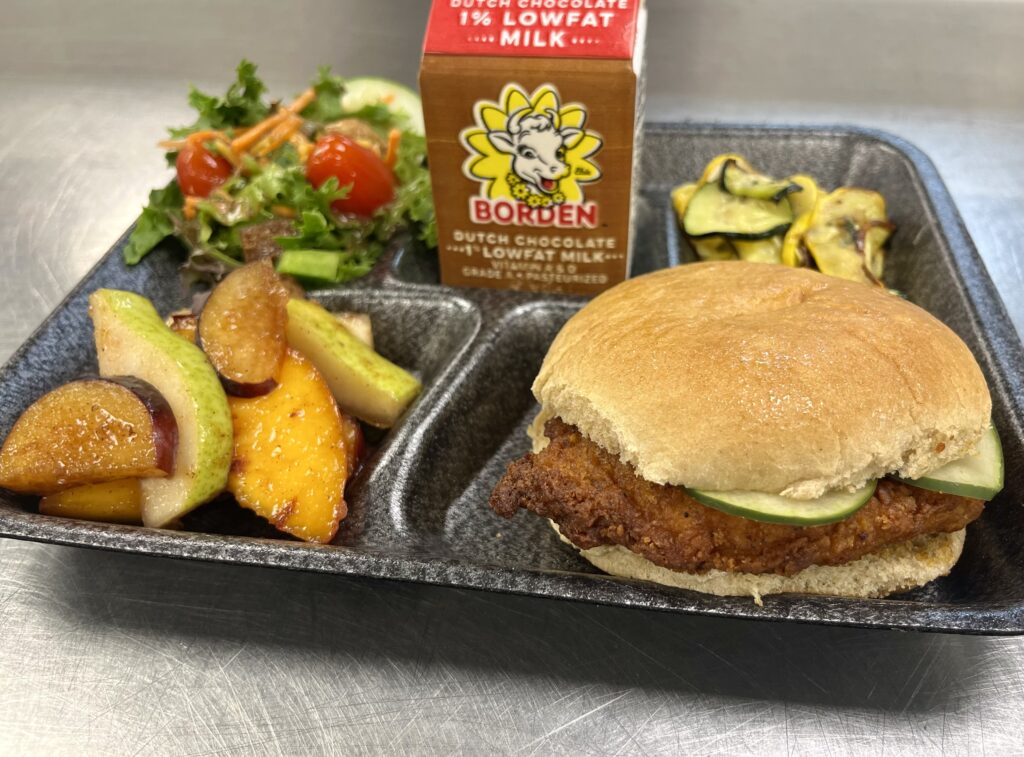 School lunch tray with salad, spiced fruit salad, squash, Nashville Hot Chicken Sandwich and chocolate milk.