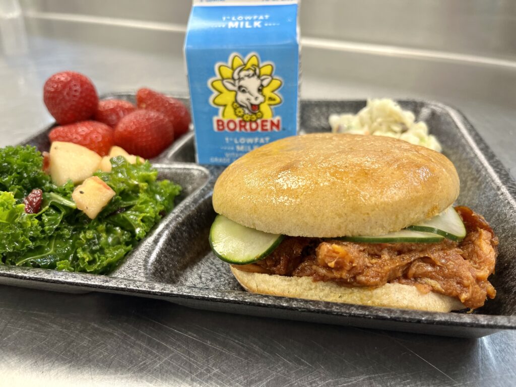 School lunch tray with BBQ Pulled Pork Sandwich, kale salad, strawberries, slaw and milk.