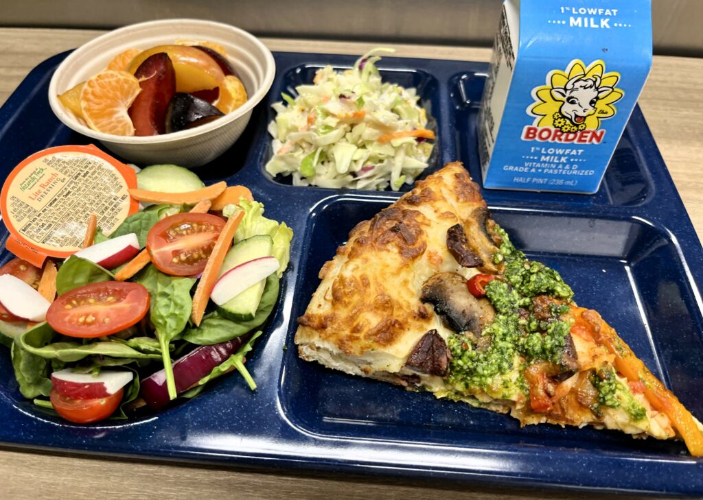 School lunch tray with Winter Greens Pesto Sauce on pizza, salad, coleslaw, fresh fruit and milk.