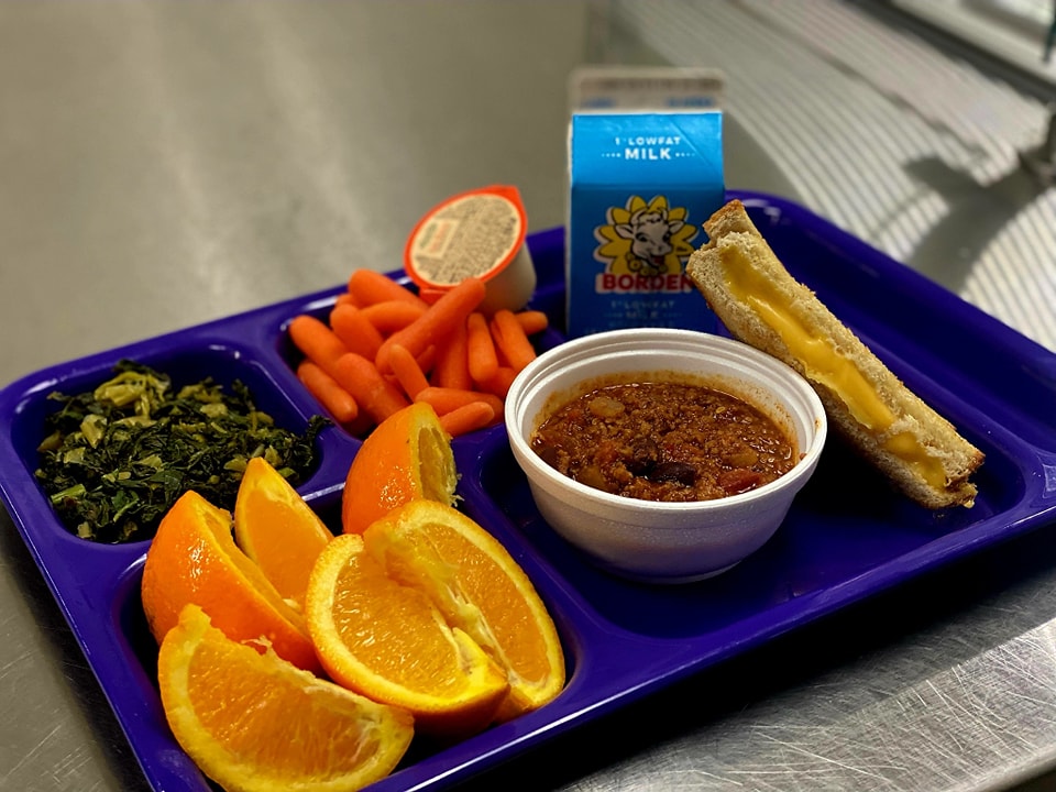 School lunch tray with chili, grilled cheese, carrots, greens, oranges, and milk.