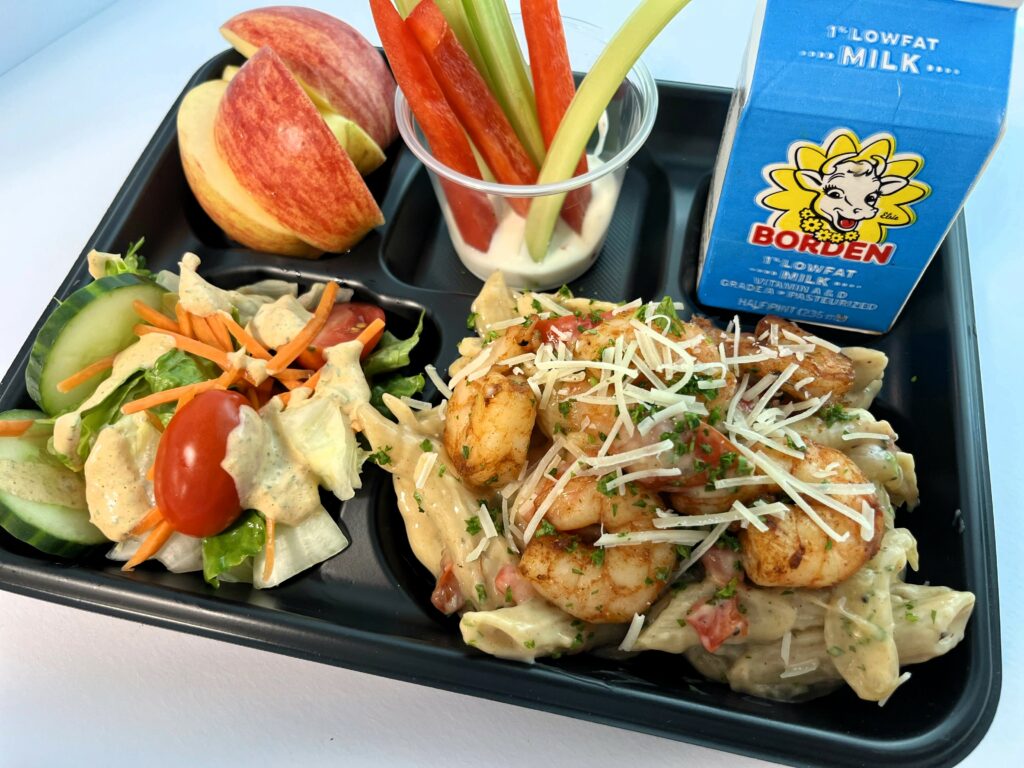 Creamy Cajun Shrimp Pasta on school lunch tray with salad, sliced apples, veggie dippers and milk.