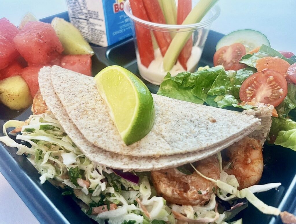 School lunch tray with Baja Shrimp Tacos, melon, veggie dippers, salad and milk.