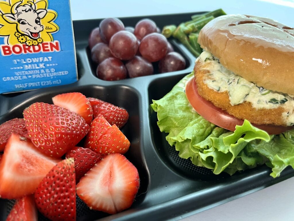 Butter Chicken Sandwich on school lunch tray with fresh strawberries, grapes, green beans and milk.