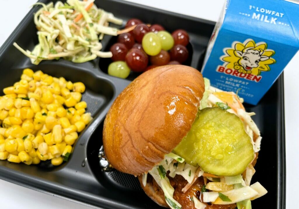 School lunch tray with Hot Honey Chicken Sandwich with Putnam Peach Slaw, grapes, steamed corn, and milk.
