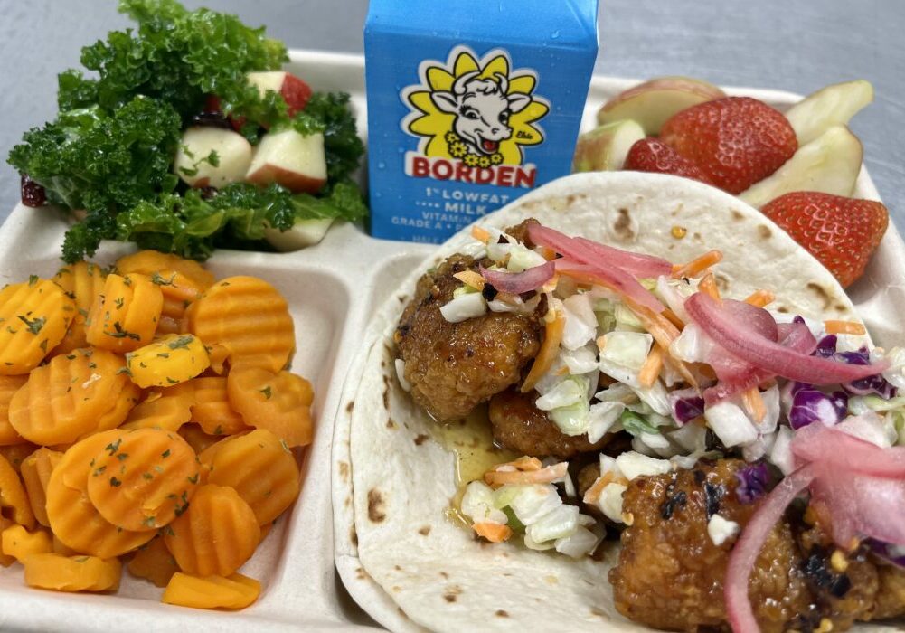 School lunch tray with Hot Orange Chicken Street Tacos, corn, kale salad, fresh fruit and milk.