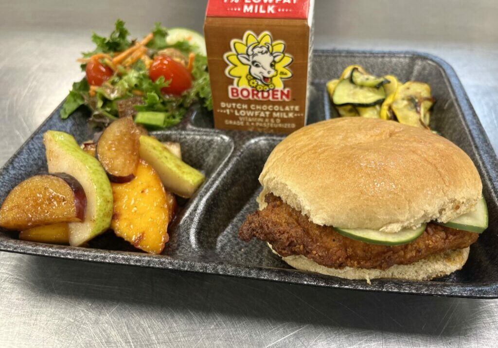 School lunch tray with salad, spiced fruit salad, squash, Nashville Hot Chicken Sandwich and chocolate milk.