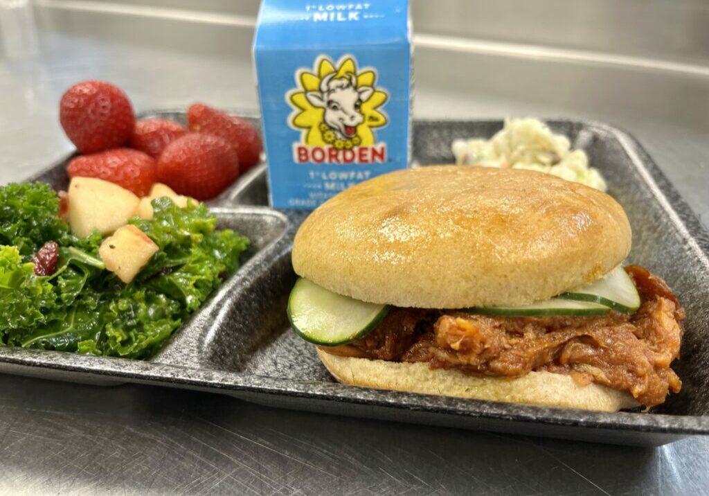 School lunch tray with BBQ Pulled Pork Sandwich, kale salad, strawberries, slaw and milk.