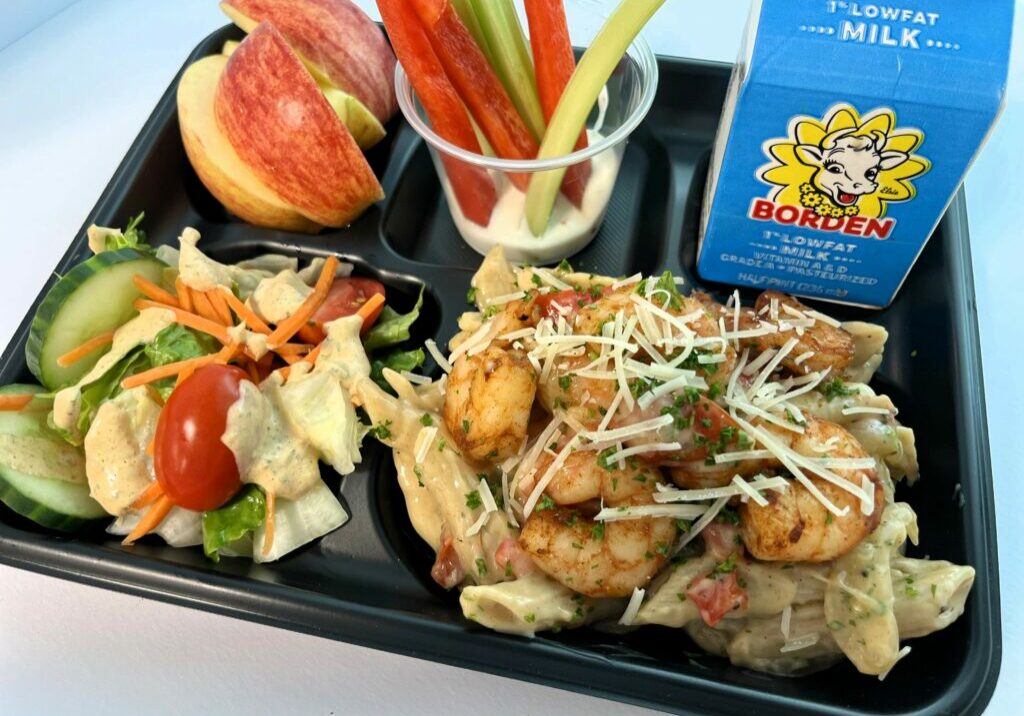 Creamy Cajun Shrimp Pasta on school lunch tray with salad, sliced apples, veggie dippers and milk.