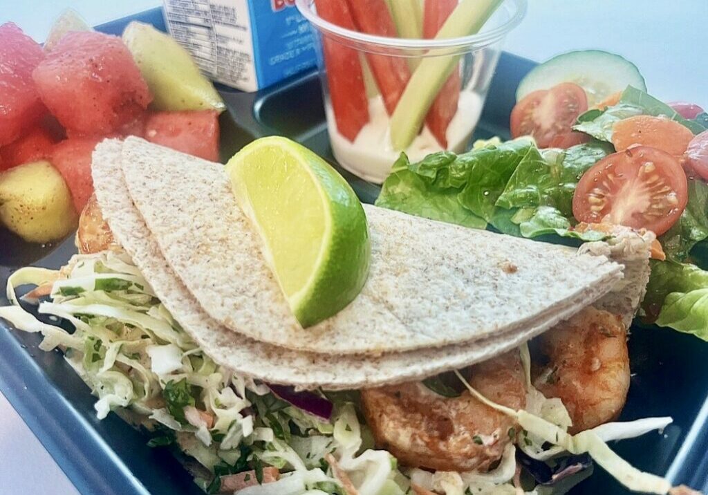 School lunch tray with Baja Shrimp Tacos, melon, veggie dippers, salad and milk.