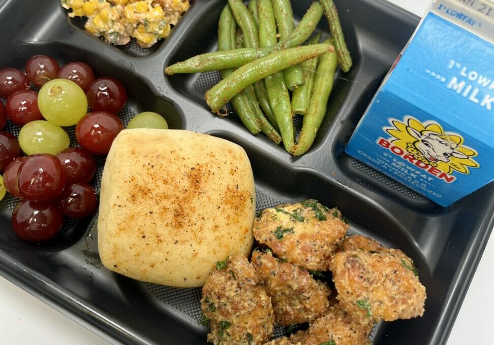 School lunch tray with Honey Lemon Pepper Boneless Wings, roll, grapes, corn, green beans, and milk.