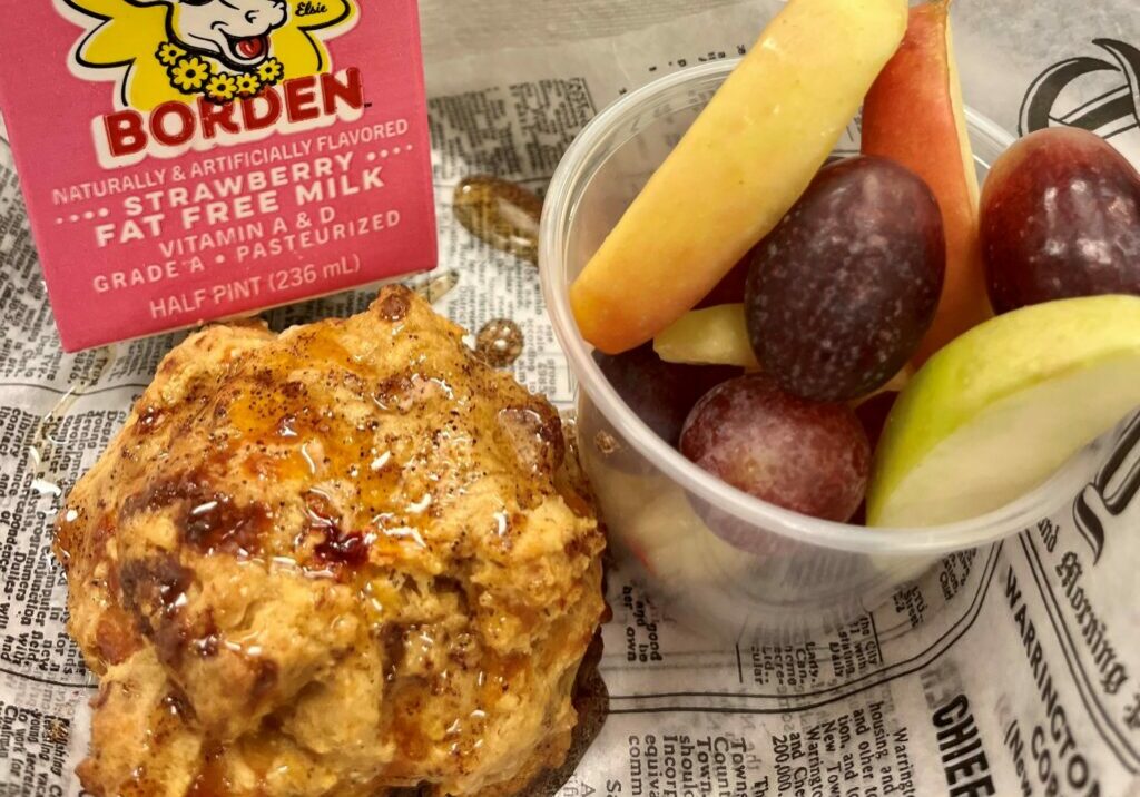School breakfast tray with Pimento Cheese Cornbread Drop Biscuit with Chipotle - Sumac Honey sauce, fresh fruit, and strawberry milk.