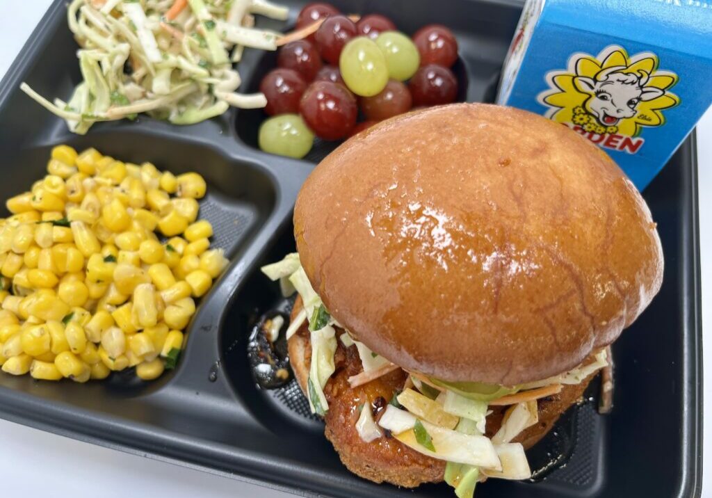 School lunch tray with chicken sandwich, slaw, grapes, steamed corn, and milk.